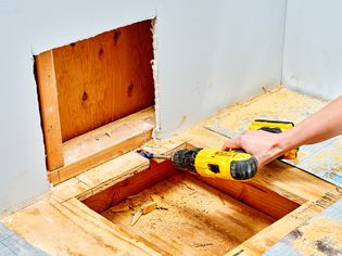 A person using a drill on a subfloor next to a wall. 